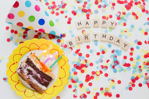 Birthday background with tasty cake on a plate, paper party hat, colorful confetti and wooden sign letters - Photo, Image