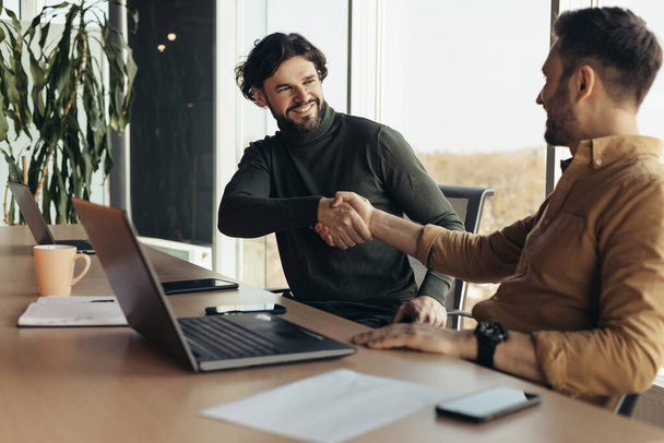 Happy business partners shaking hands in agreement, making deal, sitting at desk with laptop at modern office, copy space. Two male colleagues greeting each other, working on startup project together - Photo, Image