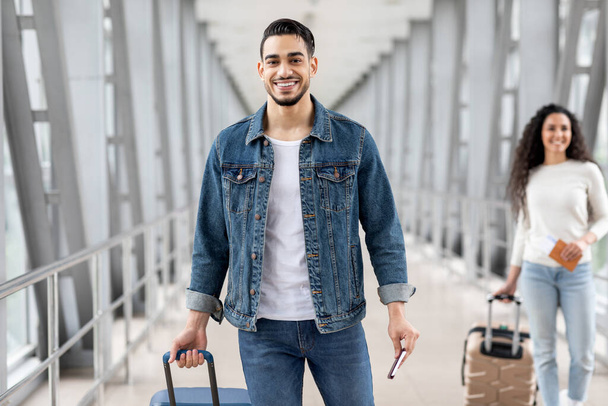 航空旅行。空港で荷物を掲げる笑顔のアラブ男、ターミナルでスーツケースを持って立つハッピー中東男性旅行者とチケットでパスポートを保持し、今後の旅行を楽しんで - 写真・画像