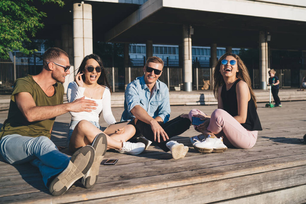Stylish friends in modern sunglasses resting and laughing together while sitting with legs crossed on street on nice warm weather - Zdjęcie, obraz