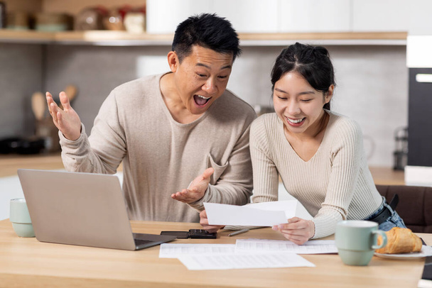 Happy asian couple paying bills online from home, sitting together at kitchen table, holding letters and papers, using computer and smiling, enjoying easy Internet banking, got good news, copy space - Photo, image