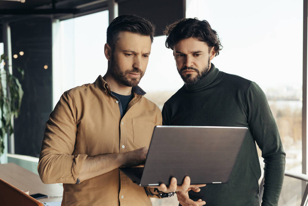 2人の深刻な男性従業員は、現代のオフィスでラップトップを使用して、共同作業、ビジネス戦略を開発しています。コンピュータの近くでアイデアや戦略をブレインストーミングする若い同僚に焦点を当て - 写真・画像