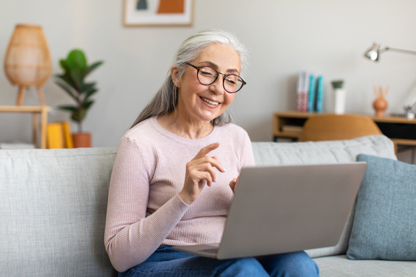 Boldog idős, ősz hajú, szemüveges nőnek van egy videohívása laptoppal, beszélgetve a közösségi oldalakon a nappaliban. Blog, munka és tanulmány távoli során covid-19 világjárvány, szabad tér - Fotó, kép