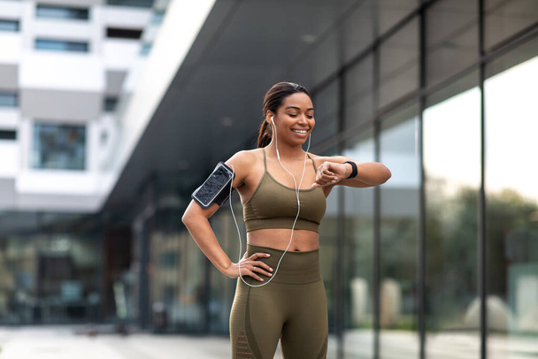 Young black woman looking at smartwatch, tracking fitness activity, listening to music in earphones during outdoor training. Fit African American female checking heart rate while jogging - Zdjęcie, obraz