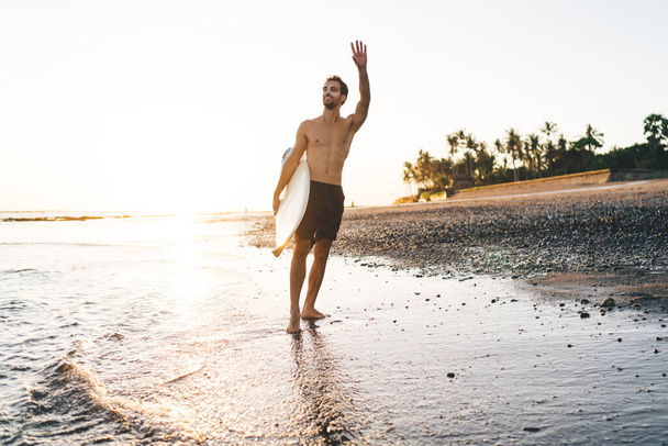 Happy shirtless male surfer in shorts with sunblock on face standing on seashore with surfing board under arm and waving hand - Фото, изображение