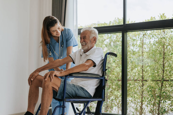 若いアジア人看護師が老人を家で助けている。老人は車椅子に座り、家の中で若い医者と話をする。シニアヘルスケア. - 写真・画像
