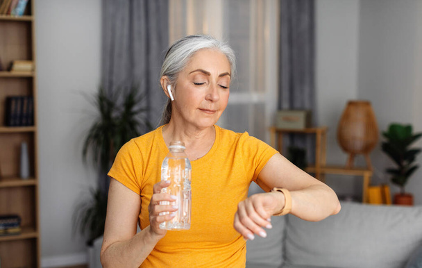 Sportowa starsza pani z butelką wody sprawdzająca zegarek lub urządzenie fitness, odpoczywająca na kanapie po treningu domowym. Starsza kobieta pozostaje nawodniona podczas treningu sportowego, prowadząc zdrowy tryb życia - Zdjęcie, obraz