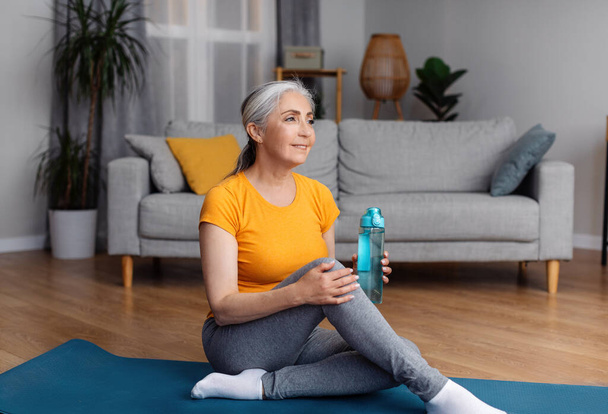 Positiv sportliche ältere Dame in Sportbekleidung sitzt mit Wasserflasche auf Fitnessmatte und lächelt, macht Pause beim Training zu Hause, Freiraum - Foto, Bild