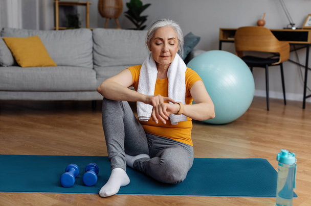 Старша спортсменка використовує смарт-годинник фітнес-трекер після домашнього тренування, перевіряючи спалені калорії, сидячи на підлозі йога килимок у інтер'єрі вітальні
 - Фото, зображення