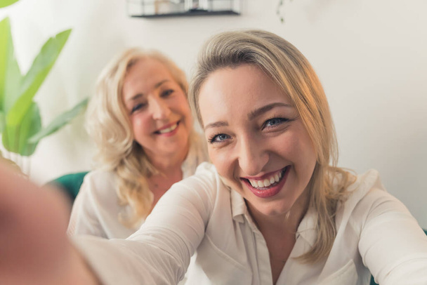Femme blonde européenne d'âge moyen en chemise blanche prenant un selfie avec sa mère au look similaire debout dans le backgruond. Coup de feu intérieur. Photo de haute qualité - Photo, image