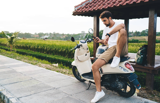 Full length of confident young male traveler sitting on scooter near near rural field and browsing smartphone during road trip in countryside - 写真・画像