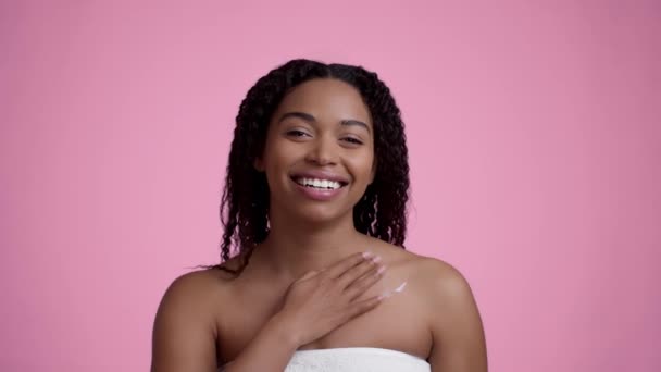Черево трупа. Закріпіть портрет молодої афроамериканки, яка посміхається, застосовуючи зволожуюче вершко на грудях після душу, рожевий фон студії, повільне переміщення - Кадри, відео