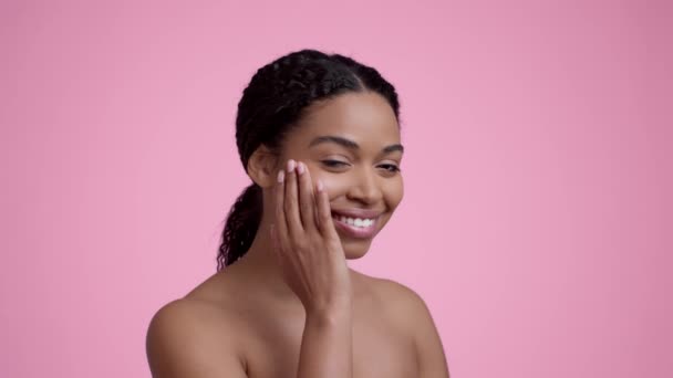 Cuidado facial. Retrato semi-perfil de una joven afroamericana bonita que aplica crema de mimos en la cara, sonriendo a la cámara sobre el fondo rosa del estudio, cámara lenta - Metraje, vídeo