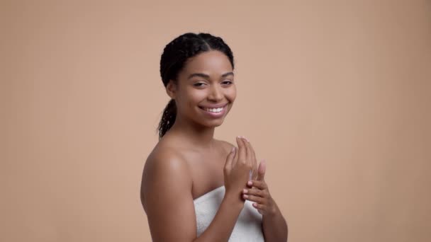 Joven mujer afroamericana positiva aplicando crema hidratante en la mano, sonriendo a la cámara, posando envuelto en toalla después del baño sobre fondo beige estudio, cámara lenta - Imágenes, Vídeo