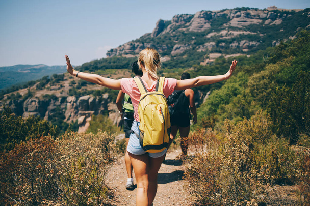 Back view of young female in casual clothes spreading arms while hiking with friends in hilly area during summer vacation - Foto, Imagen