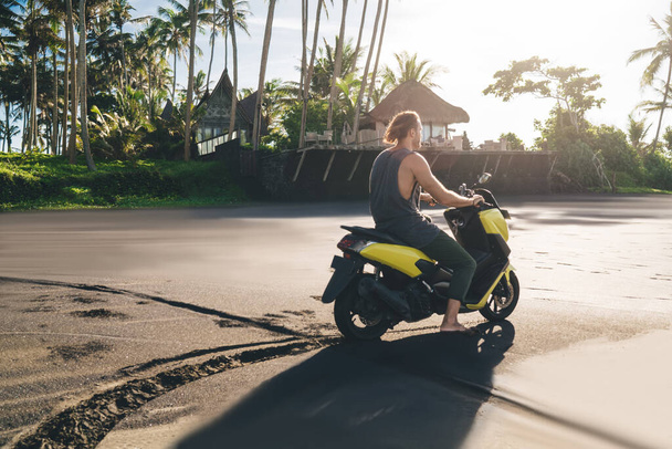 Back view of full body anonymous male driving motorbike and looking away while resting alone in tropical resort and enjoying summer vacation - Фото, изображение