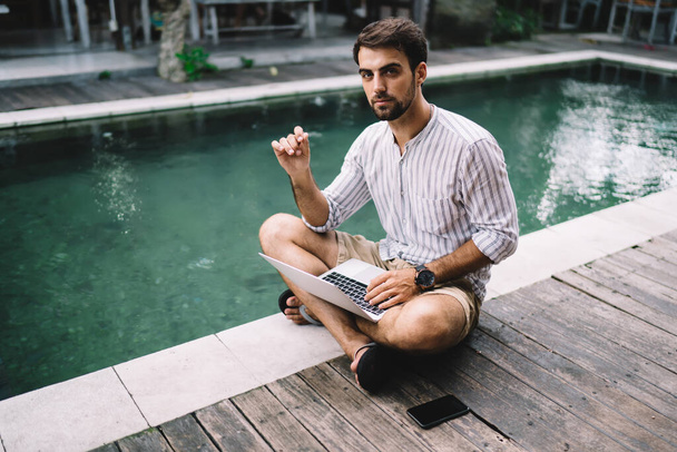 High angle of young handsome confident male freelance worker in summer shirt and shorts sitting on poolside with laptop and looking at camera - Foto, immagini