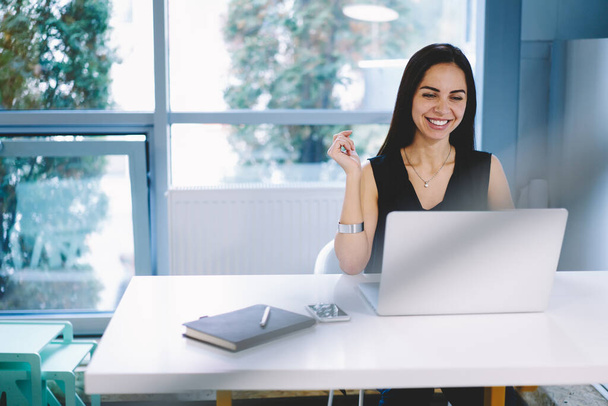 Cheerful female entrepreneur in formal clothes sitting at white table with notebook and smartphone and working on project on laptop in modern workspace - Photo, Image