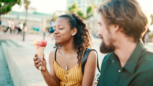 Closeup portrait of happy interracial couple eating ice cream in urban city background. Close-up of a man and woman tasting ice cream. Backlight - Photo, image