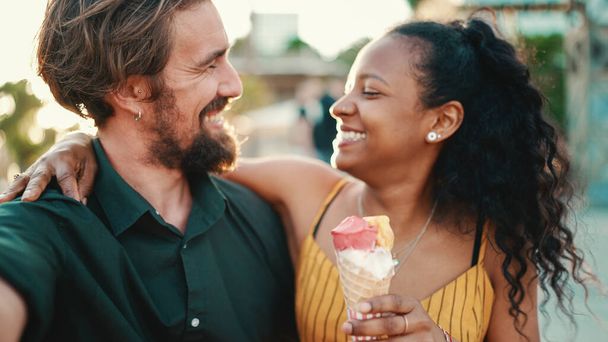 Closeup of smiling interracial couple eating ice cream and taking a selfie on urban city background. Close-up of a man and woman tasting ice cream and video chatting using a mobile phone. Backlight - Φωτογραφία, εικόνα