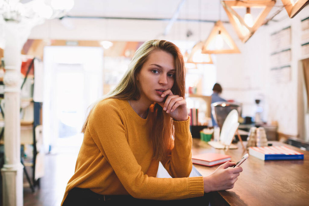 Serious woman in casual outfit touching face looking at camera sitting at wooden table browsing smartphone having rest in cafe - Photo, image