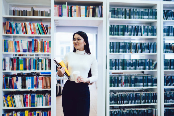 Charming Asian woman in white turtleneck with notebooks and white disposable coffee cup standing in aisle between bookshelves in light library and looking away - Zdjęcie, obraz