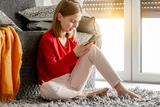 Smiling child girl communicates by smartphone and waves her hand. Cheerful kid spends time talking by video on celphone sitting on floor near window. - Fotografie, Obrázek
