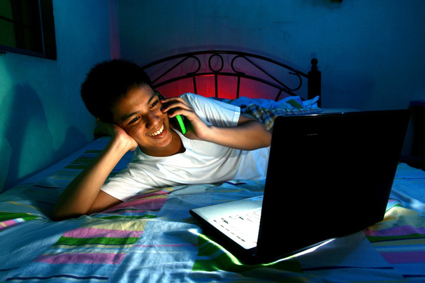 Adolescente joven frente a una computadora portátil y en una cama y usando un teléfono celular o teléfono inteligente
 - Foto, Imagen