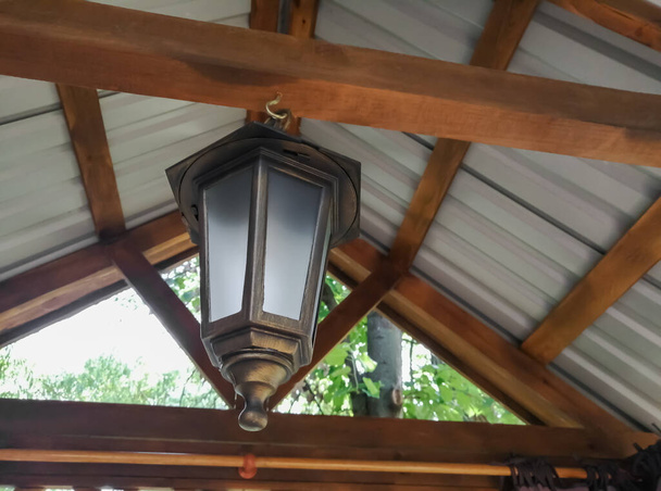 Onder een houten dak hangt een grote straatlamp. Lantaarn gemaakt van brons met matglas onder het dak bedekt met leisteen. Houten balken met straatlantaarn. - Foto, afbeelding