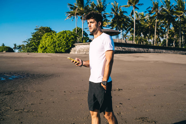 Young male athlete in sportswear with mobile phone in hand enjoying morning seascapes and looking away while standing alone on seashore - Foto, imagen