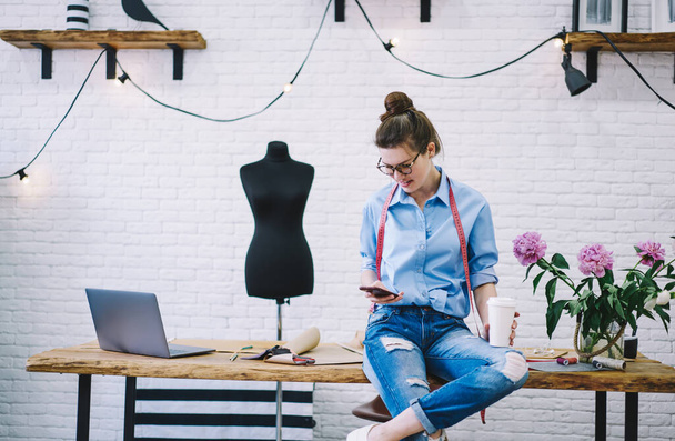 Tranquil female seamstress in glasses with takeaway hot beverage browsing smartphone and sitting crossed legs on table in creative atelier with mannequin - Foto, imagen