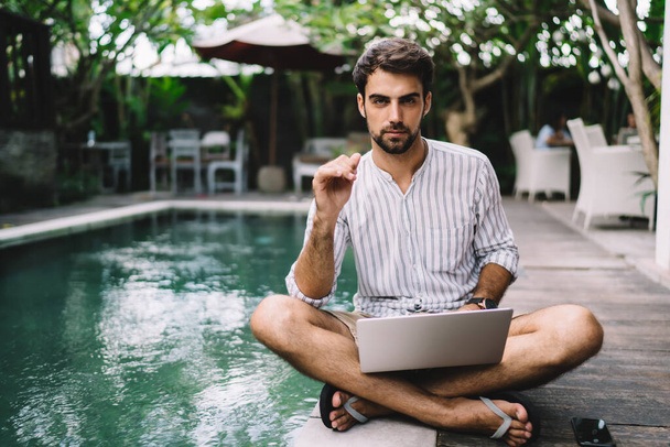 Young handsome businessman in holiday clothes sitting cross legged with laptop near empty swimming pool at wonderful spa resort and looking at camera - Photo, image