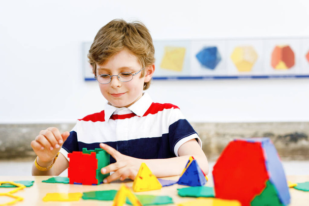 Egy szemüveges kisfiú, aki színes műanyag elemekkel játszik az iskolában vagy óvodában. Boldog gyermek építése és létrehozása geometriai alakok, tanulás matematika és geometria - Fotó, kép