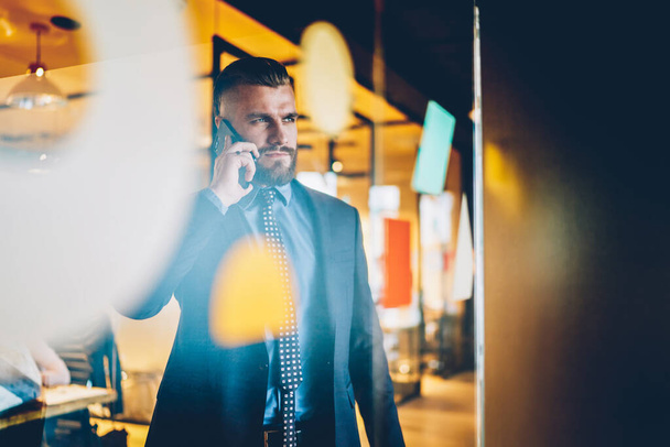 Through glass focused pleased businessman in suit talking on cellphone while looking away and standing at information glass wall in modern office - 写真・画像