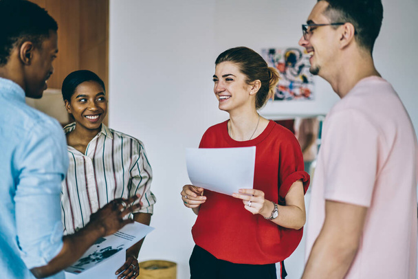 Laughing cheerful intelligent woman talking with diverse work team joyfully about company project while holding official papers during meeting at workplace - Foto, afbeelding