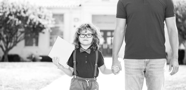 Parent and pupil of primary school go hand in hand. Teacher in t-shirt and cute schoolboy with backpack near school park - Foto, Imagen
