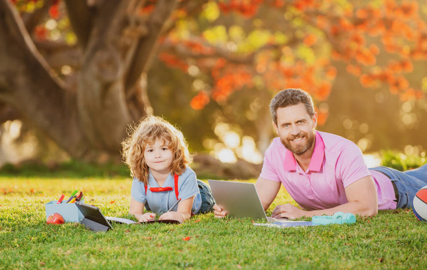 Son with father learning outdoor by studying online and working on laptop in green field - 写真・画像