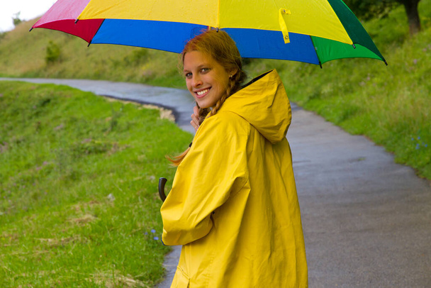 Νεαρή γυναίκα με ομπρέλα και κίτρινο αδιάβροχο περπατώντας στη βροχή - Φωτογραφία, εικόνα