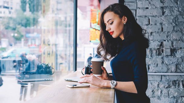 Side view of concentrated young female freelancer standing at wooden table with laptop and camera while text messaging via smartphone and drinking takeaway coffee - Photo, Image