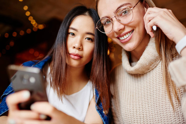 Smiling female in casual sweater with eyeglasses looking at camera and thoughtful ethnic woman with mobile phone spending time in cafe - Foto, Bild