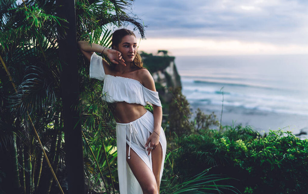 Charming young skinny woman in white elegant two piece dress leaning against poll and looking away with dreamy smile while resting near ocean - Foto, afbeelding