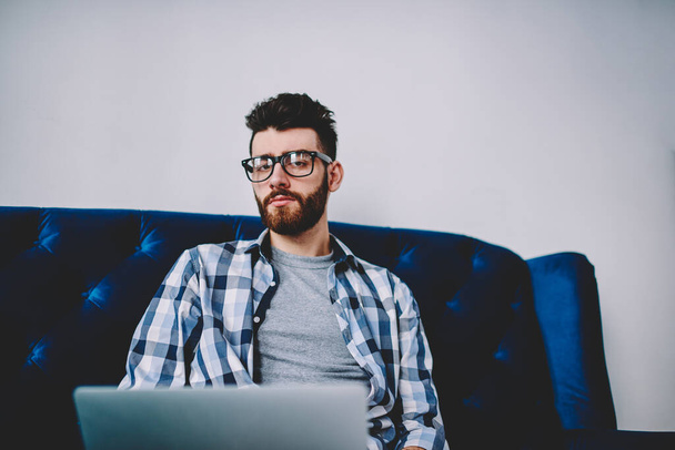 Portrait of bearded hipster guy in eyeglasses for vision correction looking at camera while working remotely ar modern laptop device using wireless internet connection.Young man sitting at computer - Zdjęcie, obraz