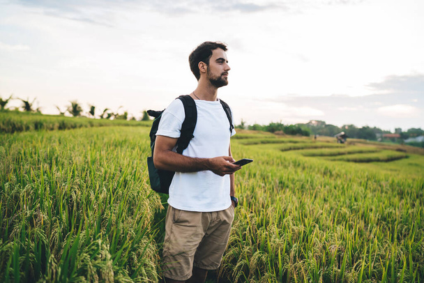 Positive young bearded male in casual outfit with backpack standing in casual summer wear using mobile phone on green vast field and looking away - Foto, Bild