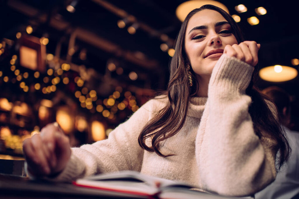 Low angle of smiling dark haired female in soft warm sweater working with notes remotely in modern cafe and looking at camera - Фото, зображення