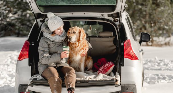 Girl sitting in car trunk with golden retriever dog in winter time with eco cup mug with hot beverage. Young woman with doggy pet labrador in auto transportation outdoors - Fotografie, Obrázek
