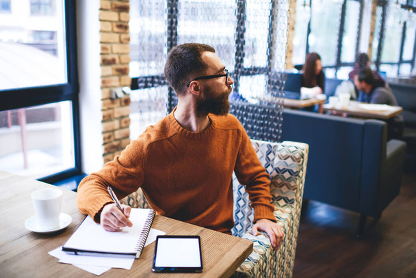 Bearded man in casual outfit and eyeglasses making order while taking notes in notebook during remote work in cafe at daytime - Фото, изображение