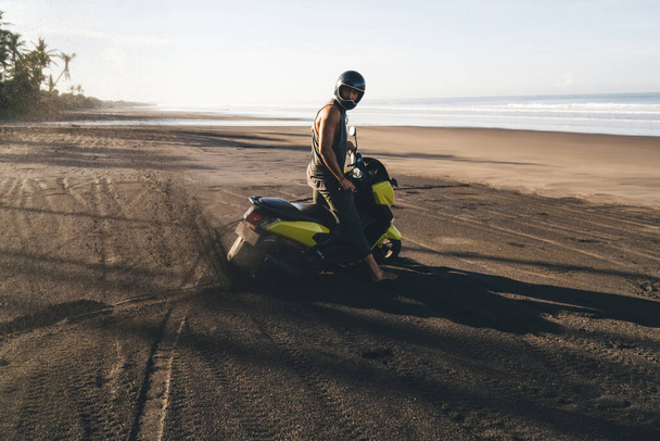 Side view full body energetic male motorcyclist in helmet standing near motorbike on black sandy seashore and looking at camera on sunny weather - Foto, Imagem