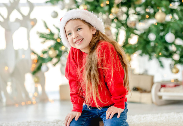 Dziewczynka dziecko w kapeluszu Mikołaja w czasie Bożego Narodzenia siedzi i uśmiecha się na urządzone Nowy Rok domu z choinką. Cute dziecko w czasie Bożego Narodzenia - Zdjęcie, obraz