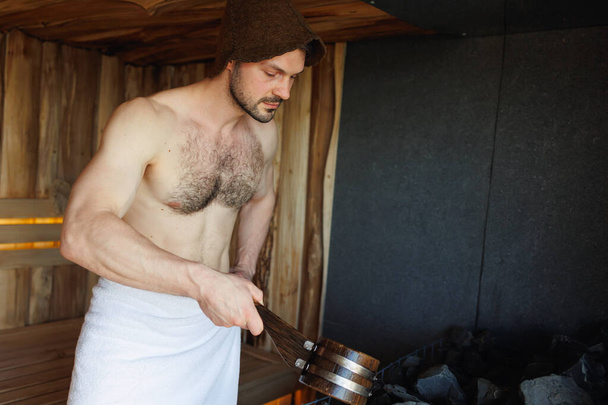 Brutaali mies saunassa kaatamalla vettä kauha kuumille kiville - Valokuva, kuva