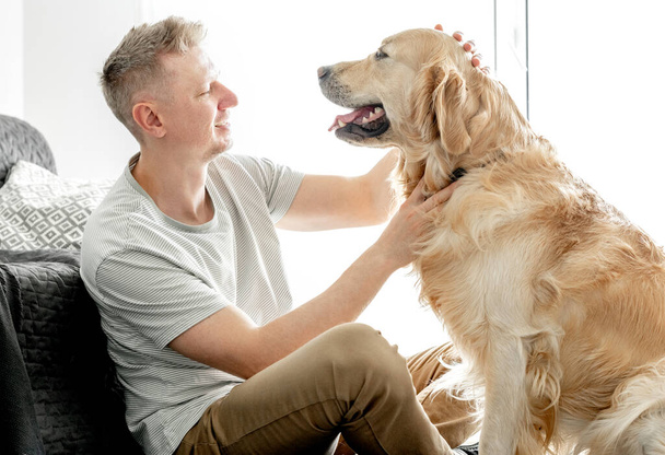 Young handsome man with his golden retriever dog in bright living room. - Foto, Imagem
